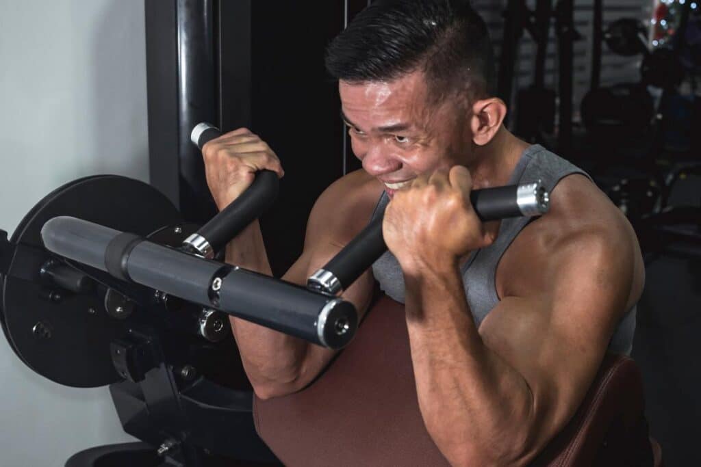 A buff asian male does some machine bicep curl preacher at the gym. Arm and bicep workout training. Fitness in men at their 40s