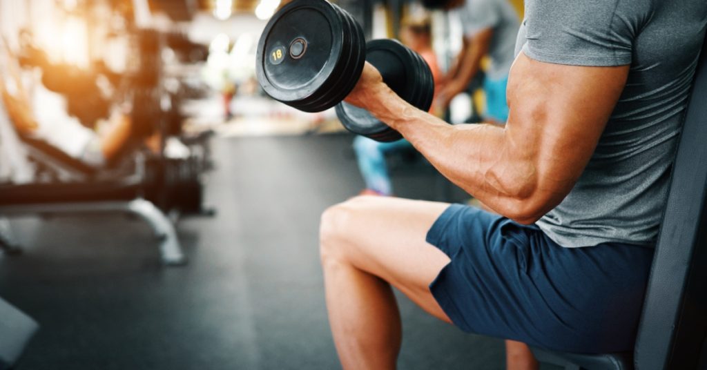 A man in a gym lifts a dumbbell with his left hand while sitting on a chair. He is wearing a gray shirt and blue shorts.