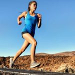 A woman in shorts and a blue tank top sprinting down a black asphalt road in the desert, her arms pumping.