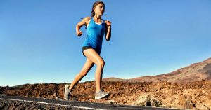 A woman in shorts and a blue tank top sprinting down a black asphalt road in the desert, her arms pumping.