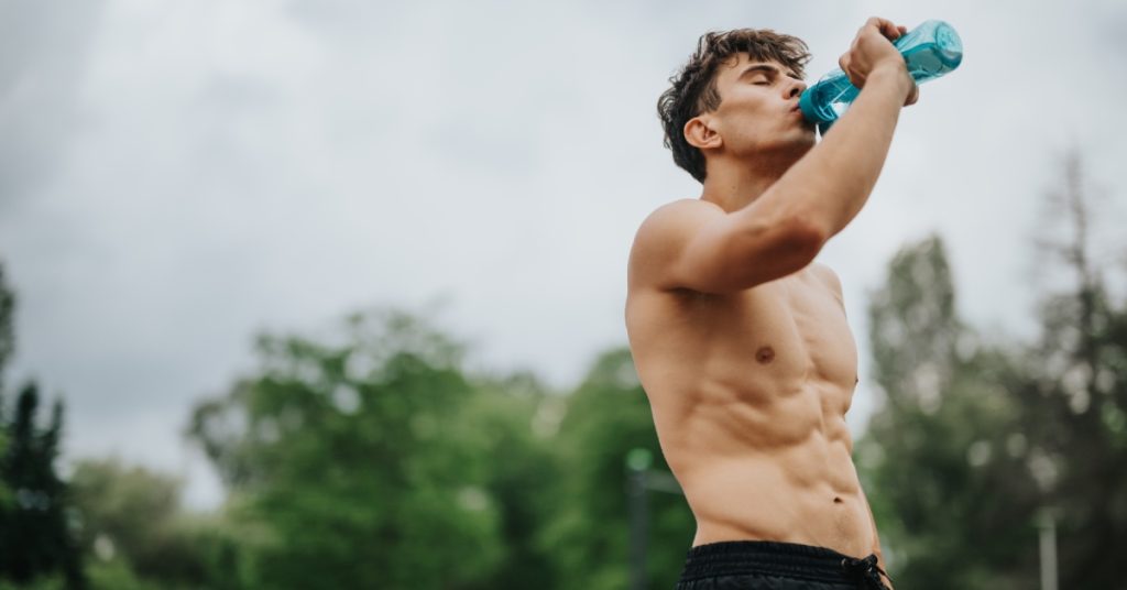 A young man is shirtless and drinking water from a teal reusable water bottle. He is wearing black workout shorts.