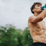 A young man is shirtless and drinking water from a teal reusable water bottle. He is wearing black workout shorts.