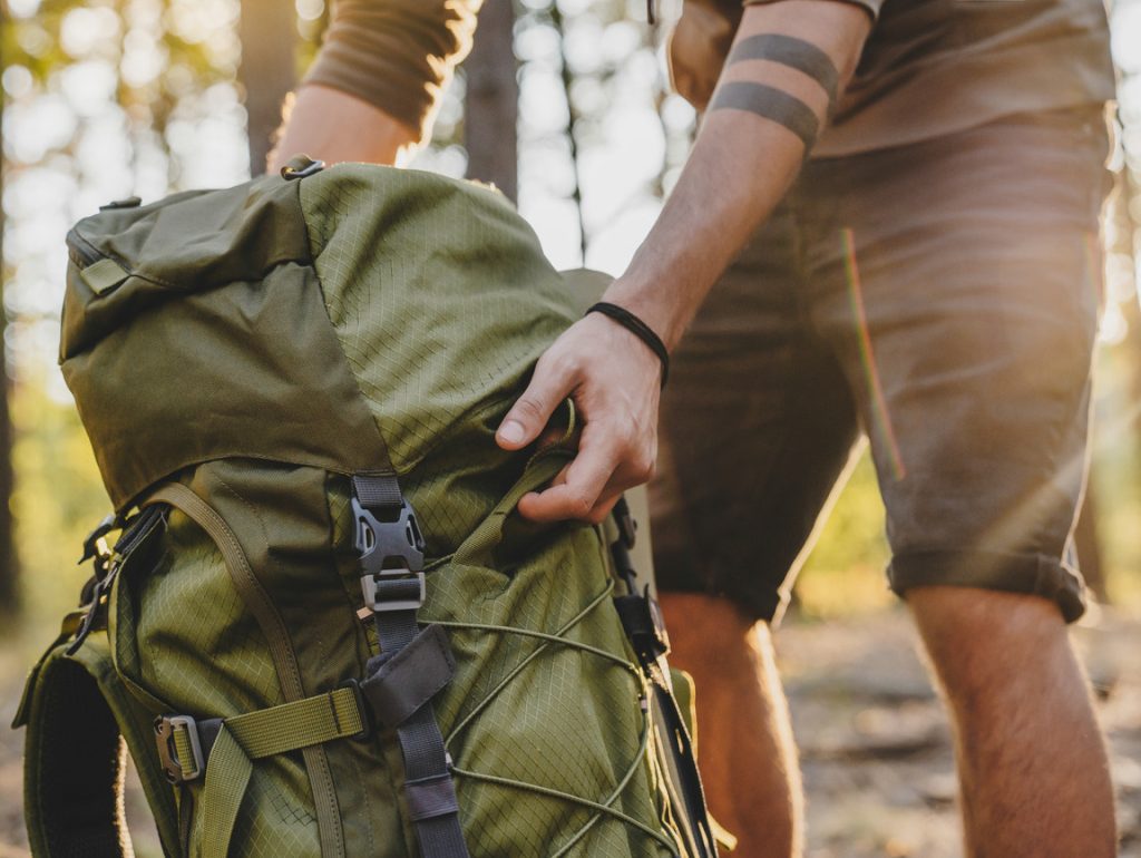 A backpacker in a forest bending down to lift his muted green tactile backpack by a pair of straps on its sides.