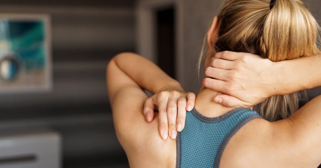 The back of a woman wearing a sports bra and her hair in a ponytail. She is using both hands to massage her neck and shoulder.