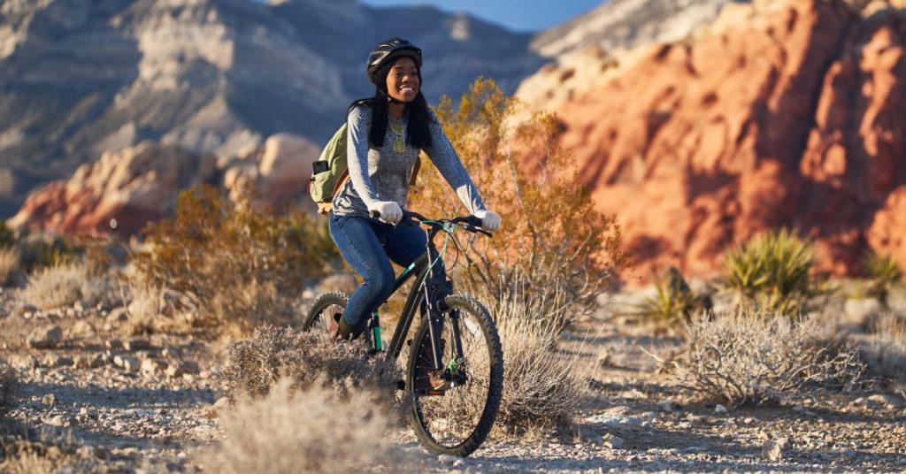 A young black woman rides her bike through a canyon in the desert. She is smiling and wearing a protective helmet.