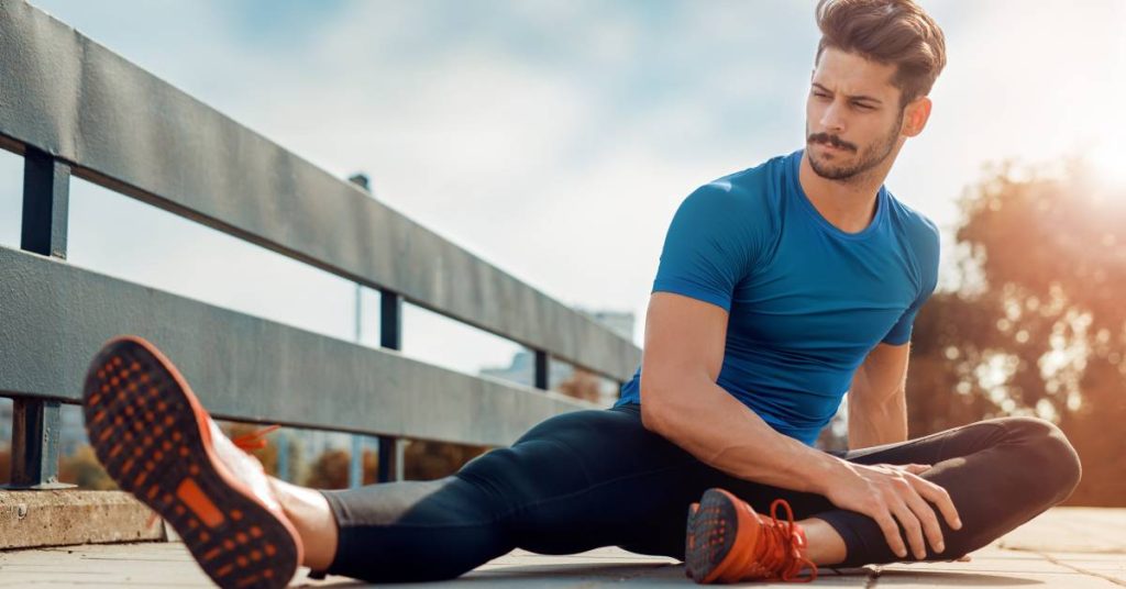 A male runner on a wooden walking bridge who is sitting on the ground stretching his legs before a run.