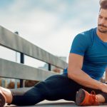 A male runner on a wooden walking bridge who is sitting on the ground stretching his legs before a run.