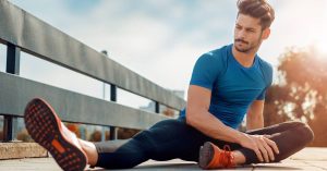 A male runner on a wooden walking bridge who is sitting on the ground stretching his legs before a run.