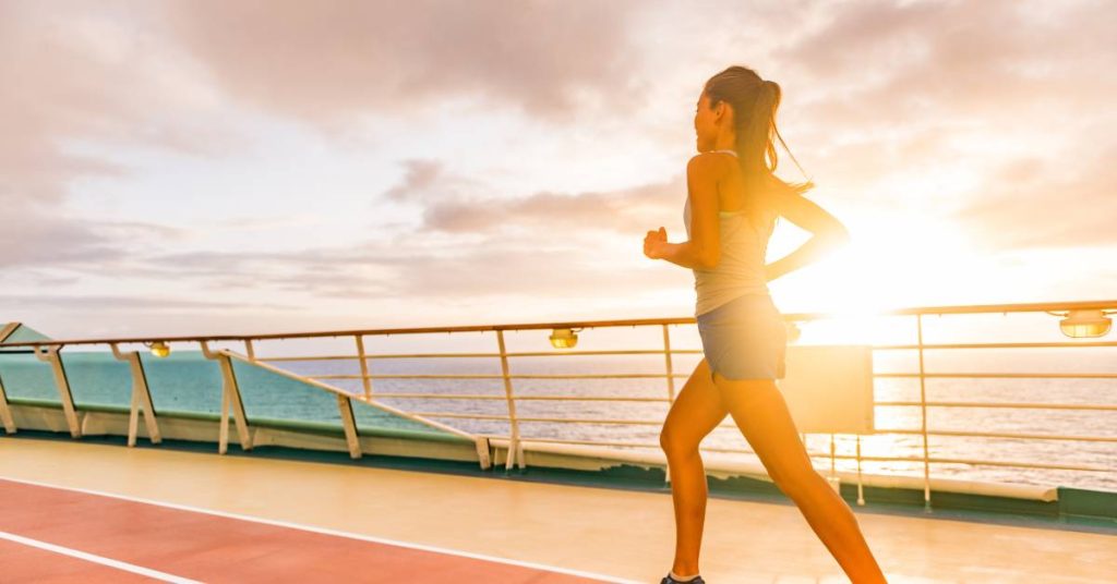 A woman wearing black shorts and shoes runs on a cruise ship deck. The sun is setting in the background.