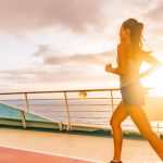 A woman wearing black shorts and shoes runs on a cruise ship deck. The sun is setting in the background.