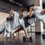 A father and son in a gym in push-up positions with one arm holding themselves up as they lift dumbbells with the other.