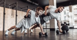 A father and son in a gym in push-up positions with one arm holding themselves up as they lift dumbbells with the other.