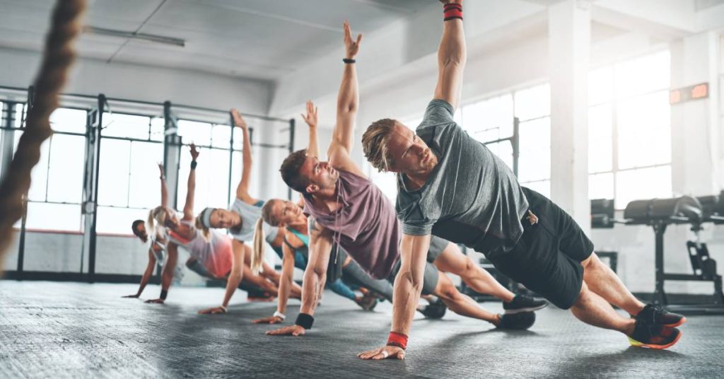 A group of six individuals in a gym engaging in the same balance exercise, extending one arm up and balancing on the other.