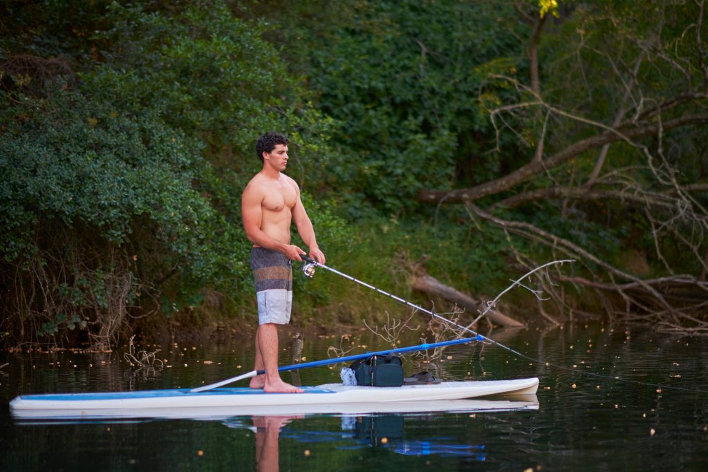 A young man fishing as he stands on a paddleboard. He's wearing swimming trunks and the fishing line is in the water.