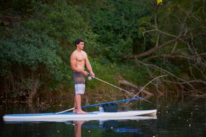 A young man fishing as he stands on a paddleboard. He's wearing swimming trunks and the fishing line is in the water.