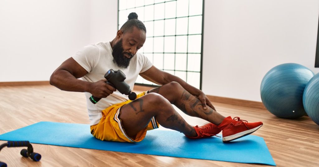 A fit man with tattooed calves sitting on a stretching mat and holding an activated percussion massage tool to his thigh.