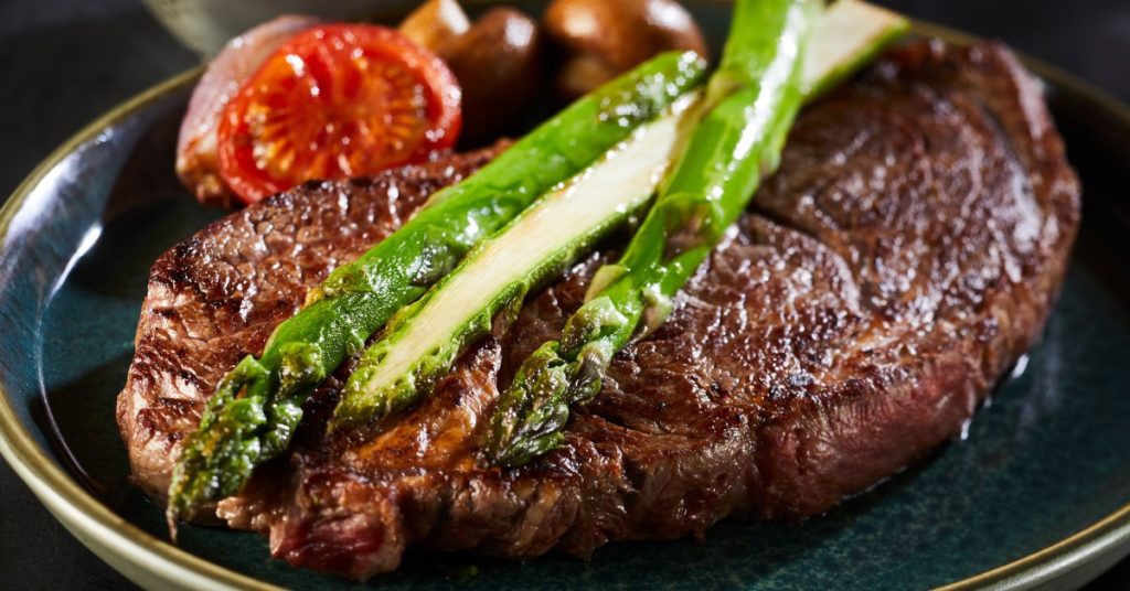 A juicy sirloin steak topped with roasted asparagus and accompanied by tomatoes and mushrooms on a dark blue plate.