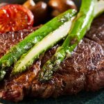 A juicy sirloin steak topped with roasted asparagus and accompanied by tomatoes and mushrooms on a dark blue plate.