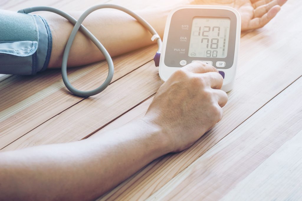 A person sitting down at the table to monitor their blood pressure. They're using a cuff attached to a digital reader.