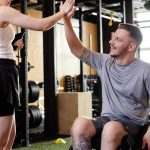 A coach and her client, who is using a wheelchair, high fiving each other. They ae both in a gym with fake grass.