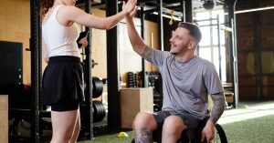 A coach and her client, who is using a wheelchair, high fiving each other. They ae both in a gym with fake grass.