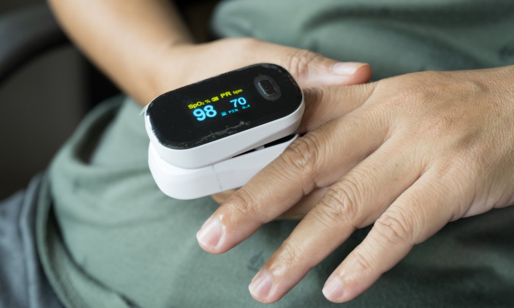 A person resting in a chair while a pulse oximeter sits around their index finger. Its digital screen has results on it.