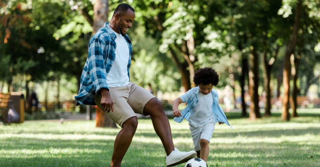 A Black father and son happily playing soccer in the backyard. The dad is going in for a kick as the son chases the ball.