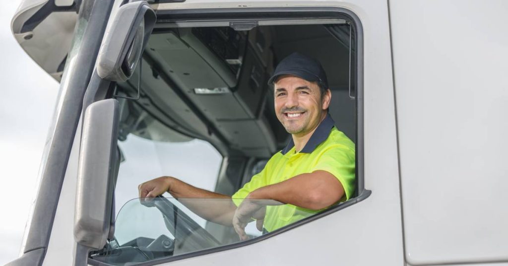 A man in a navy hat and a yellow polo shirt with a navy collar smiles as he looks through a truck window.