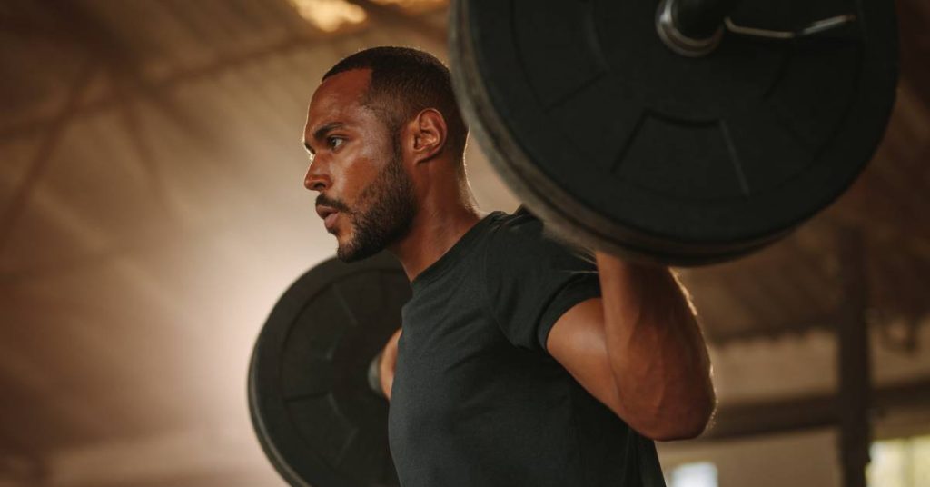 A close-up of a man in a black T-shirt lifting a barbell with large weights on each end above his shoulders.