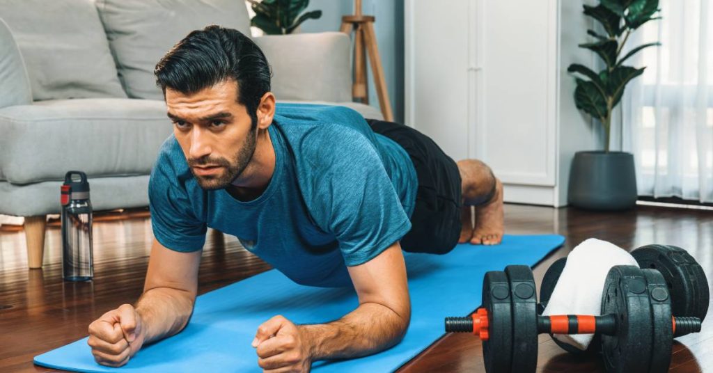 A bearded man wearing a blue T-shirt does a plank exercise on a blue yoga mat next to a set of weights.