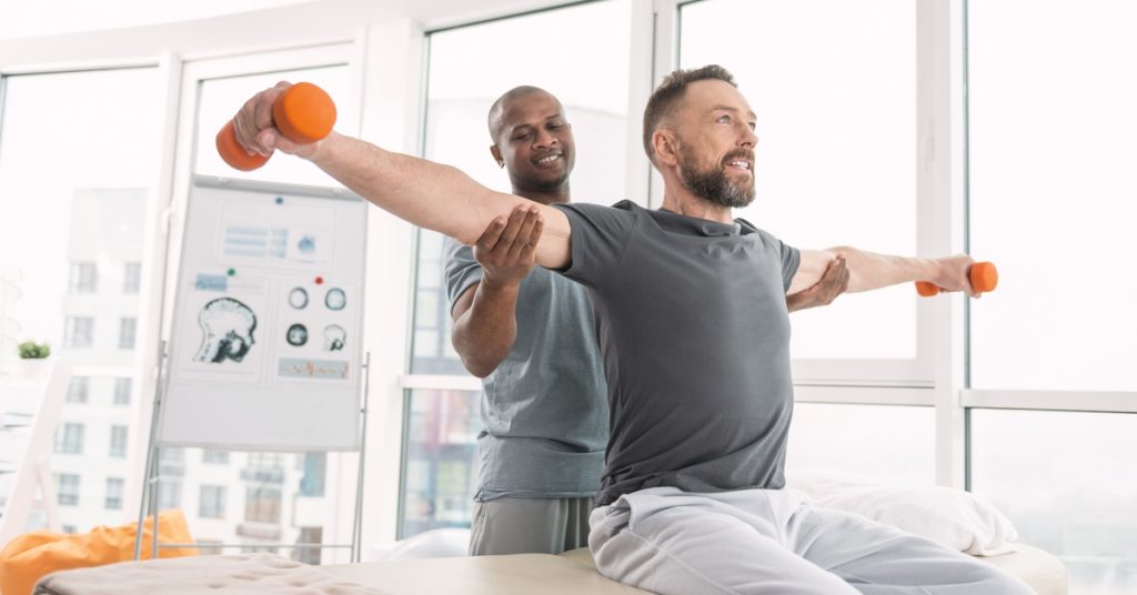 A man holding orange dumbbells with his arms stretched out to his side during a physical therapy session.